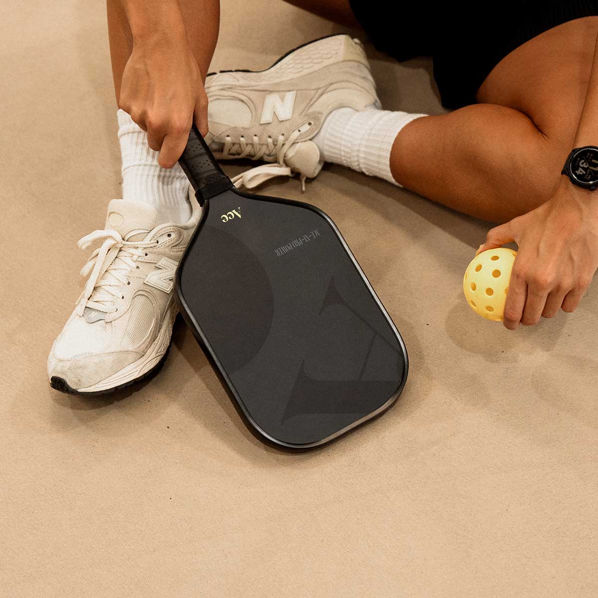Person sitting on a beige surface with a black paddle and yellow ball, wearing white sneakers.