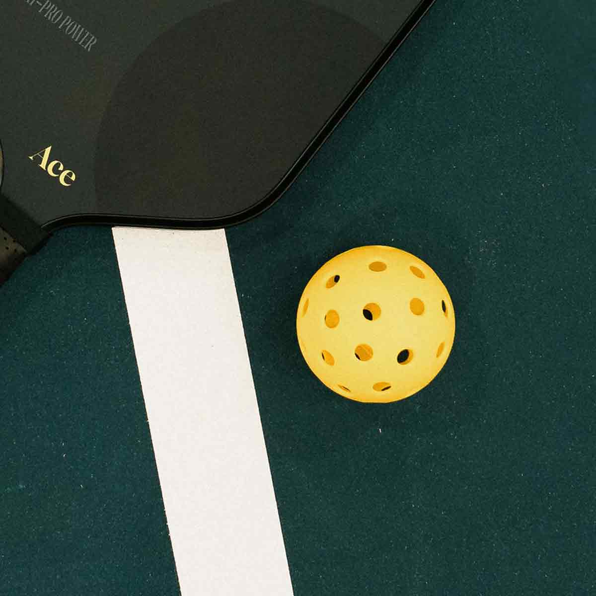 Yellow pickleball on a green court with a black paddle labeled 'Ace'.
