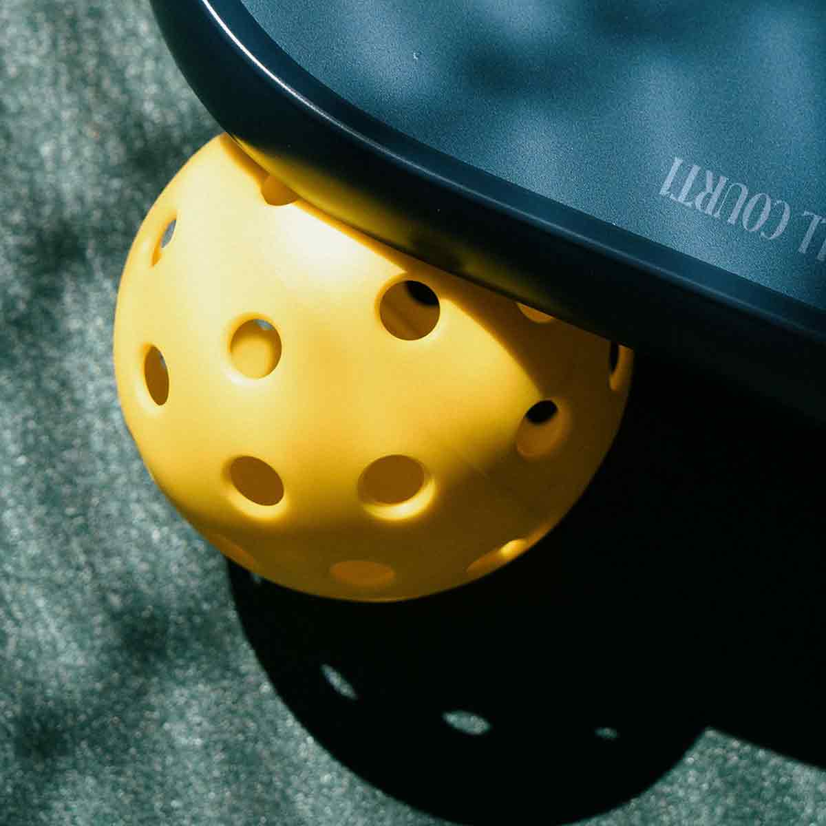Yellow perforated pickleball on a green surface with a blue paddle.