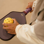 Person holding a pickleball paddle and yellow pickleball against a beige background