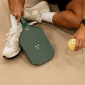 Person holding a green pickleball paddle and yellow pickleball on a beige surface