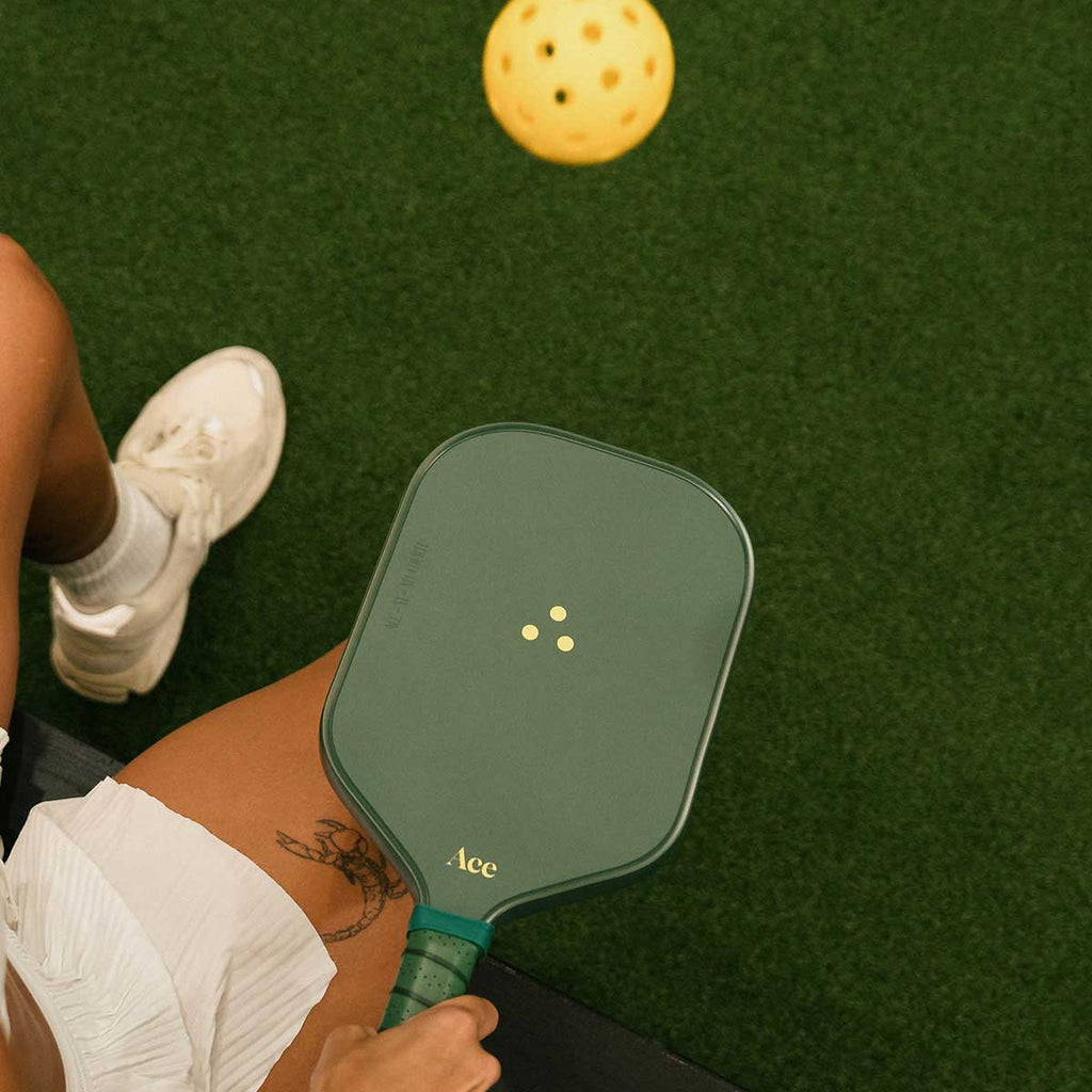 Person holding a green pickleball paddle on a green court with a yellow pickleball above.
